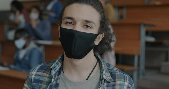 Close-up portrait of young biracial man wearing medical face mask standing in high school class. Corona virus pandemic and education concept.