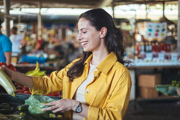 Young woman buying fresh organic produce at a local farmer's market. Picking vegetables at local green market. Young woman on the market. Cheerful woman selecting fresh vegetables in market