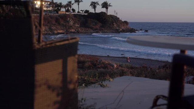 Wide shot Mediterranean sea coast with woman picking stuff on pebble beach at golden sunset. Blurred armchair and table on pier at front. Carefree Caucasian tourist at sundown on Cyprus