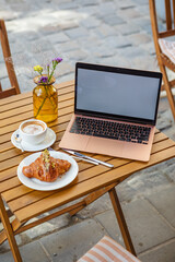 a laptop with a white screen, a croissant with coffee on a table on the summer terrace in a cafe