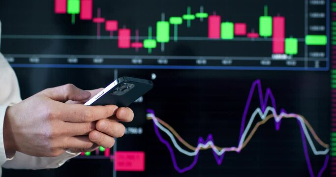 Influencer in work clothes holds phone against dynamic rate chart on plasma screen. Businessman types message about working moments at workplace in office closeup