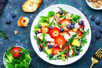 Healthy summer salad with strawberry, grilled chicken slices, fresh herbs with blueberries, avocado and walnuts, blue stone table., top view