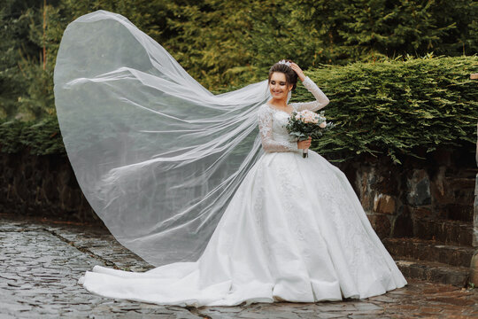 Full length side view of one beautiful sensual young brunette bride in long white wedding dress and veil standing in park holding bouquet outdoors on natural background, horizontal picture