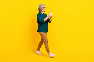 Full size profile portrait of positive lady use smart phone walking isolated on yellow color background