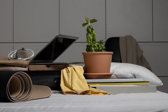 Clothes, laptop in a suitcase with plant, charger, books, pillow and a yoga mat  prepared on the bed. Behind it is a chair with chlotes on it.