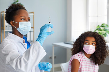 Vaccination. African American female doctor prepares syringe with vaccine for immunization of teenage girl. Dark-skinned preteen girl watches as nurse in medical mask releases air from syringe.