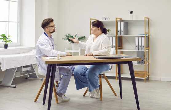 Portrait Of A Young Male Friendly Doctor Listening And Giving Consultation To A Young Fat Overweight Woman Patient Sitting At The Desk In Office During Medical Examination In Clinic.