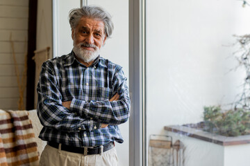 A gray-haired man in a shirt stands against the background of a large window. A smiling, gray-haired man with a beard stands idly in the house.