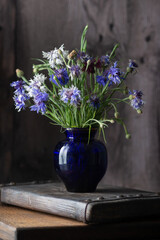 Still life with cornflowers in a blue glass vase and wooden boards on a table. Dark and moody style - 613157941