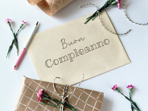 Envelope and greeting card with fresh carnation flowers. Wrapped gift, paper postcard, envelope, cord, scissors and purple carnations. Text buon compleanno means happy birthday in Italian language.