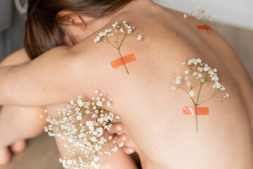 Back view of young naked slim woman embracing knees with twigs of white flowers of gypsophila stuck...