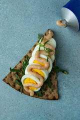 Crisp bread open sandwich with boiled egg, cod roe paste and fresh arugula on light blue background.