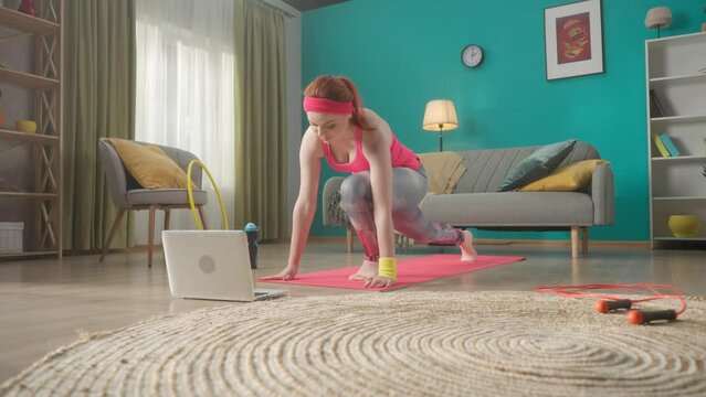 Redhaired woman in sportswear doing a warmup, stretching on a sports mat in the living room. A woman is watching a video tutorial on a laptop. Home fitness. Slow motion. HDR BT2020 HLG Material.