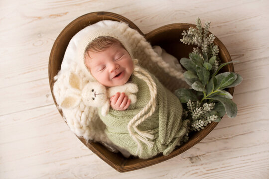 A cute newborn baby in a green pistachio winding and a white hat smiles sweetly and sleeps. Wooden basket in the shape of a heart. Knitted white rabbit in a pen. photo against a light wooden floor.