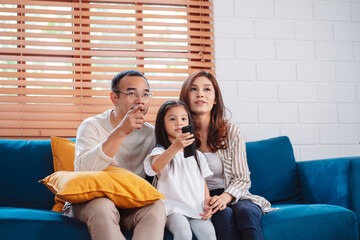 Asian family consisting of happy parents and daughter watching TV or movie together on sofa in living room at home. enjoy relaxing happiness