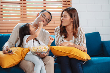 Asian Family consisting of parents, happy daughter watching TV or movie eating popcorn on sofa in...