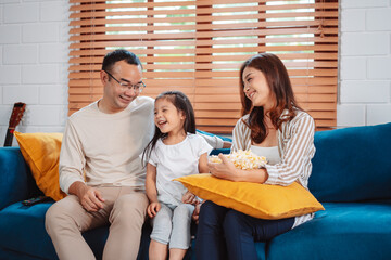 Asian Family consisting of parents, happy daughter watching TV or movie eating popcorn on sofa in living room at home. enjoy relaxing happiness.