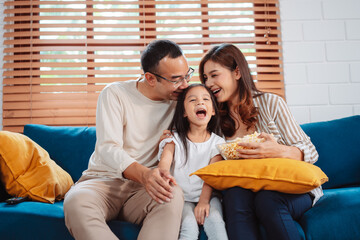Asian Family consisting of parents, happy daughter watching TV or movie eating popcorn on sofa in living room at home. enjoy relaxing happiness.