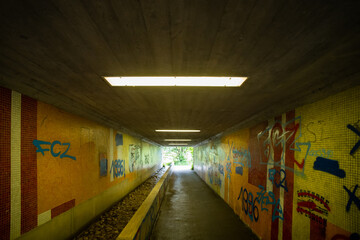 Neon lit urban pedestrian tunnel with graffiti tile side walls and small running water stream, no people