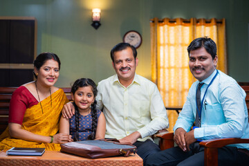 Happy indian banker looking camera in front of middle class family with kid at home looking at...