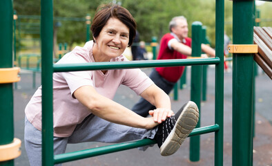Positive elderly woman doing leg stretching on outdoor sports ground