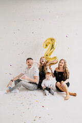 Mom, dad, daughter congratulate son on birthday near white wall in studio. Happy family having fun celebrating birthday party 2 years old. Confetti. Open cracker confetti, falling and flying glitter.