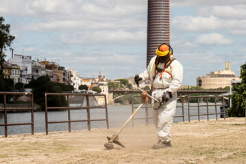 man clearing dry grass with a brush cutter dressed in a safety suit and mask. outside a city.