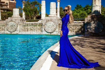 Glamour, stylish elegant woman in blue long evening gown dress is posing near the pool in luxury...
