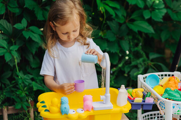 Child washes toy dishes and playing with toy kitchen outdoors in summer. Play area including...
