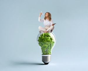 Idea of renewable energy and energy saving. Woman with good news is sitting on a light bulb filled with leaves. - 613200374