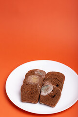 Rotten moldy sliced rye brown bread on white round ceramic plate from above on bright orange background. Spoiled bakery. Unhealthy food.