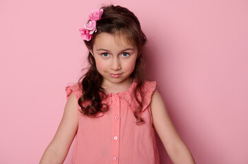 Close-up portrait face of a beautiful Caucasian little girl 5-6 years old, with blue eyes and stylish hairstyle, dressed in elegant pink dress, looking at camera, isolated over pink color background