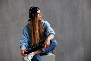A young pretty long haired DJ girl in a blue sweater, jeans and a black baseball cap poses while sitting with a black DJ mixing console on her lap. Studio shot, gray background.
