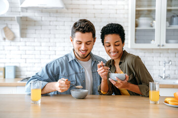 Healthy breakfast. Weekend morning. Happy young family couple, caucasian man and african american woman, having breakfast at cozy home kitchen with healthy porridge, juice and fruits, smiling