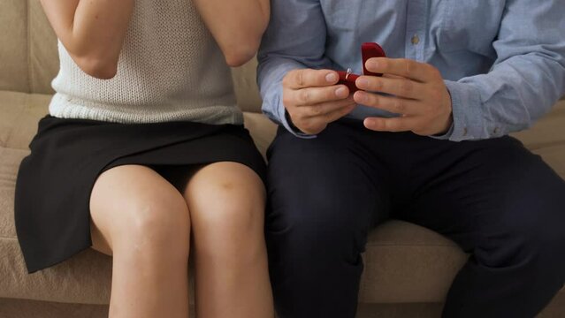 A Caucasian man and woman are sitting on a sofa in the living room of the house. He takes out a red box with a gold ring, puts it on her finger and makes a marriage proposal. She agrees