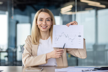 Portrait of a young business woman sitting in the office and showing documents with graphs to the...