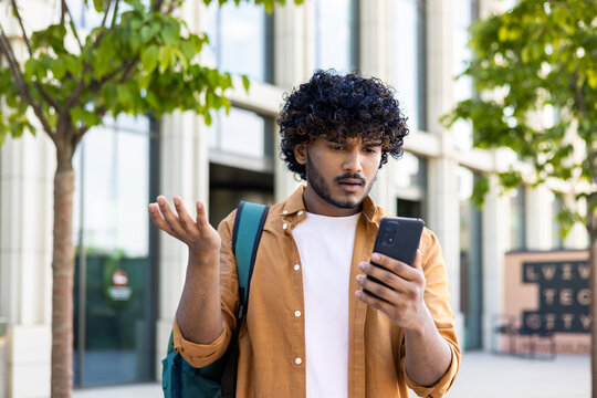 Upset student with backpack reads bad news on phone, man with phone browses internet page on smartphone, opens app, hispanic outside near university campus