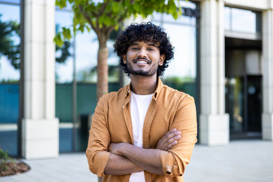 Portrait of young hispanic freelancer businessman outside office building, successful man looking at camera and smiling and with wrinkled hands, worker with curly hair and wearing a shirt