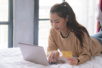 An asian woman in brown sweater using labtop and credit card for online shopping at home with smile of happiness. Girl holding credit card while laying down and using laptop on bed with smile.