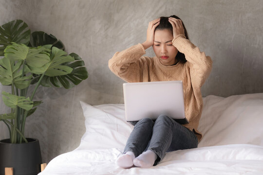An asian woman in brown sweater and jean put hands over head and fold eye bow together while working on bed, freelance girl got stress while using computer.