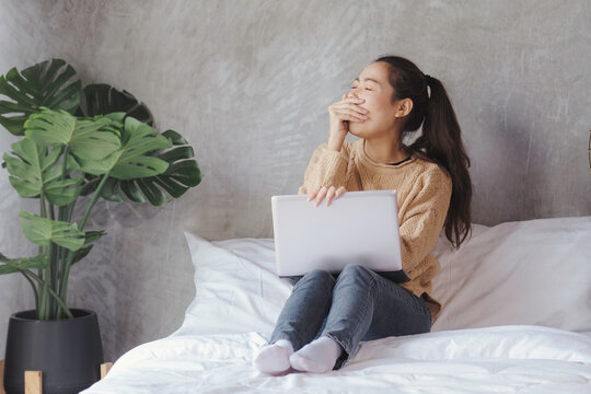 Business girl in brown sweater using labtop, working on bed, student yawning while learning online at home.