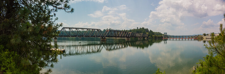 Panorama of metal train bridge over river with a dam and reflections on the water