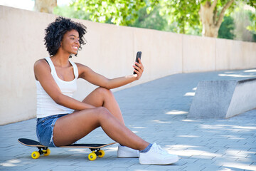 smiling african american young girl sitting on top of her skateboard taking a selfie