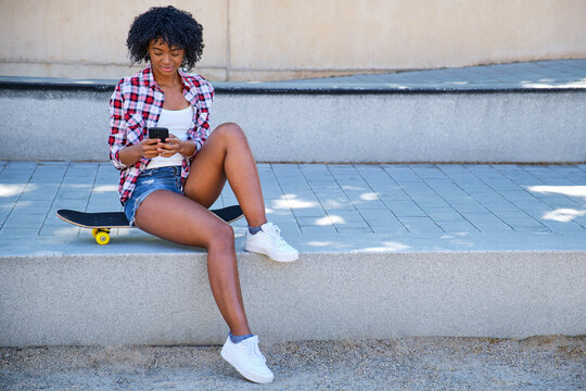 young afro american girl sitting on a skateboard looking at her smart phone