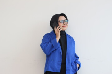 portrait of beautiful asian woman in hijab, glasses and wearing blue blazer making phone call and smiling