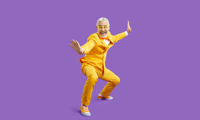 Fototapeta na wymiar Funny senior man in yellow suit pretends like he knows martial arts. Happy cheerful crazy bearded guy in funky outfit posing in kung fu fighting stance and smiling isolated on solid purple background
