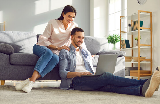 Young married couple sitting on sofa in the living room at home using tablet PC for internet and social media. Happy family resting on couch enjoying weekend watching video or talking on video call.