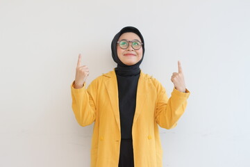 Beautiful young Asian Muslim woman in glasses, hijab and wearing yellow blazer smiling confident and happy with both hands pointing up