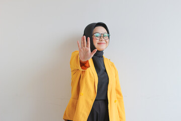 Beautiful young Asian Muslim woman, wearing glasses and yellow blazer showing stop gesture while smiling