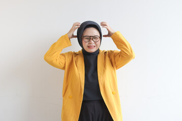 Beautiful young Asian Muslim woman, wearing glasses and yellow blazer showing stress gesture while holding head